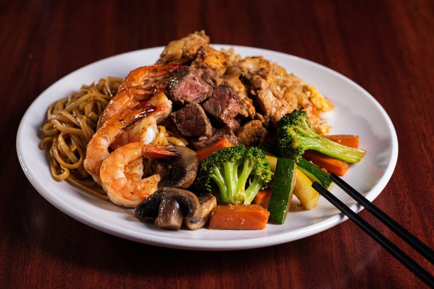 A white plate of hibachi steak, shrimp and chicken with noodles, broccoli, zucchini, carrots and mushrooms, chopsticks across the rim